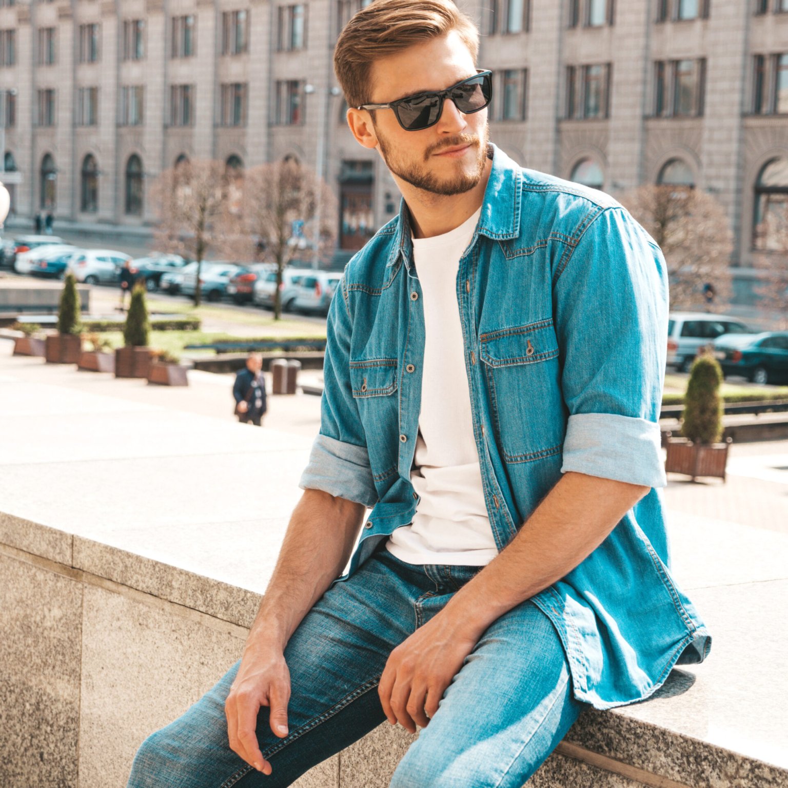 Portrait of handsome stylish hipster lumbersexual businessman model. Man dressed in jeans jacket clothes. Fashion male posing on the street background in sunglasses